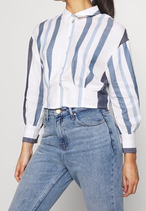 Camicetta corta a maniche corte in bianco con righe verticali blu e navy, chiusura con bottoni e colletto a punta; abbinata a jeans blu a vita alta.