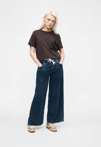 Γυναίκα με ξανθά μαλλιά που φοράει μαύρο t-shirt, φαρδιά μπλε τζιν παντελόνια με λευκή κορδονένια ζώνη και μπεζ αθλητικά παπούτσια με animal print.