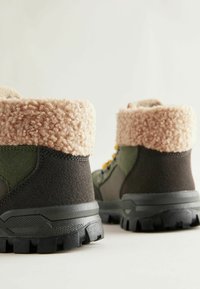 Wysokie buty w ciemnozielonym i czarnym zamszu z pluszowym beżowym kołnierzem z sztucznego futra, z chropowatą gumową podeszwą i żółtymi akcentami sznurowadeł.