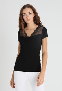 Schwarzes, figurbetontes T-Shirt mit kurzen Ärmeln und Spitzenüberzug am V-Ausschnitt. Hergestellt aus weichem Stoff und kombiniert mit weißen Hosen.