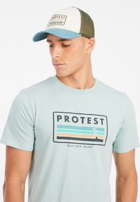 Šviesiai mėlyna trumpomis rankovėmis marškinėliai su grafiniu "PROTEST" logotipu. Žalia tinklinė krovininė kepurė su kontrastinga mėlyna skyle ir panašiu prekių ženklu.