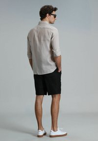 Chemise en lin beige clair, manches retroussées, shorts noirs, et baskets blanches avec semelles brunes ; vue de dos sur un fond neutre.