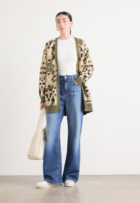 Zaļi un bēši adīts kardigans ar ģeometriskām rakstām, savienots ar augstas jostas maksiem ziliem džinsiem un baltām sporta apaviem. Bēšs somas kabatas.