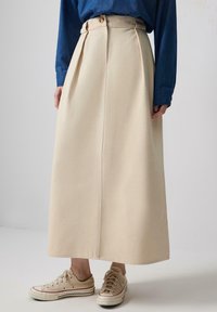 Beige, knee-length skirt with pleats, high waist, and a button detail at the top. Paired with light beige canvas sneakers.