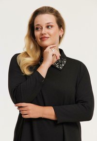 Schwarzes Kleid mit einem Kragen, der mit verzierten Details versehen ist. Lange Ärmel, weiches Material und lockere Passform. Das Model posiert mit einer Hand am Kinn.