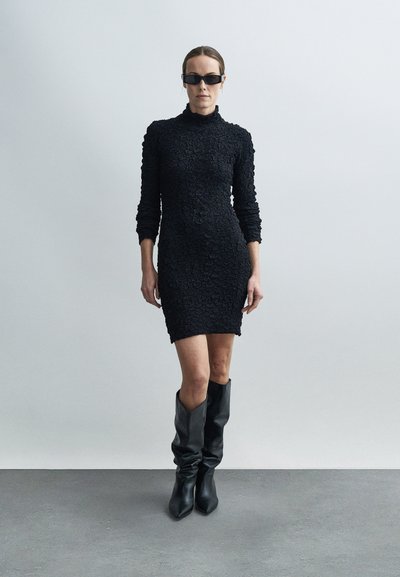 Naine, kes kannab tekstuuriga musta pika varrukaga minikleit, mustasid laiade säärega põlvikuid ja mustaid ristkülikukujulisi päikeseprille, seisab hallil põrandal.