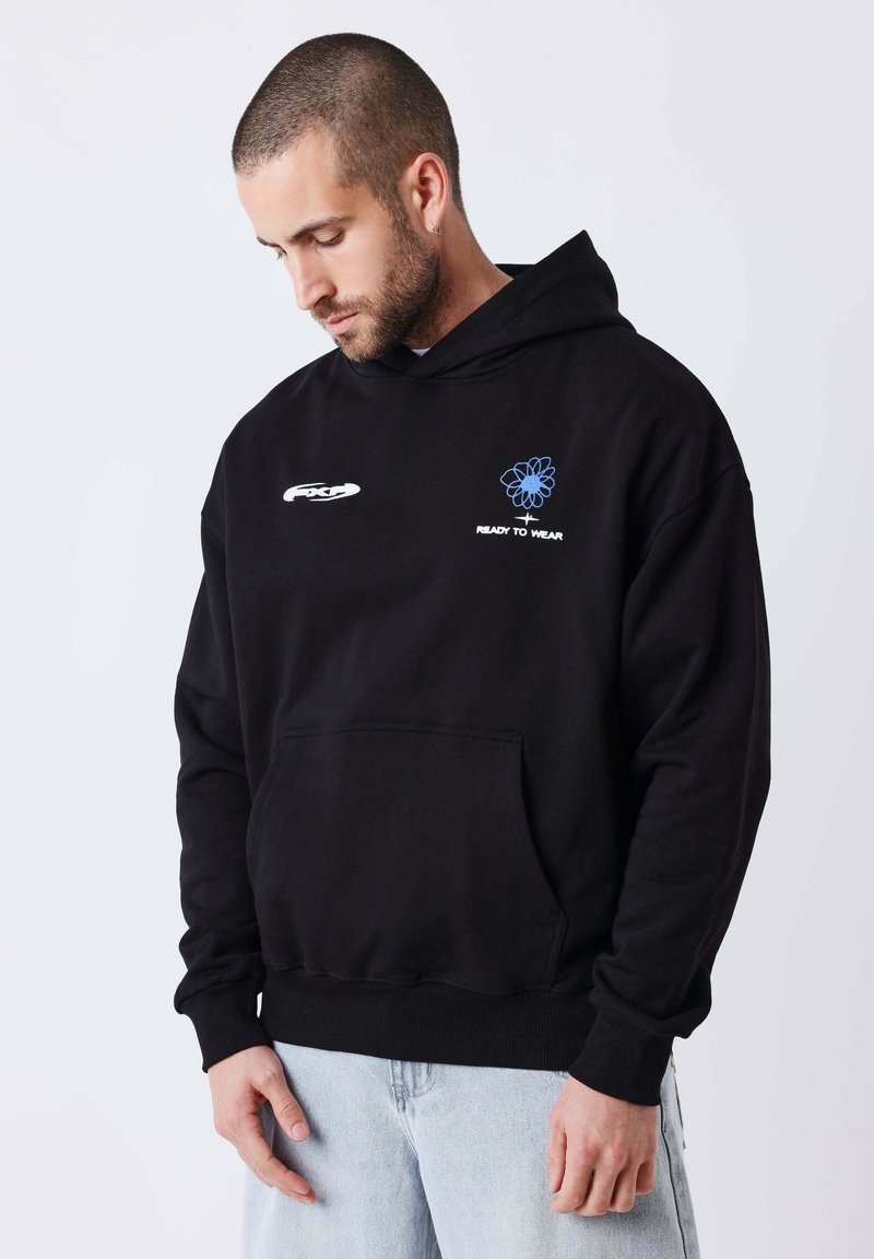 Jeune homme regardant vers le bas, portant un sweat à capuche noir avec une fleur bleue et le texte « Ready to Wear », associé à un jean bleu clair.