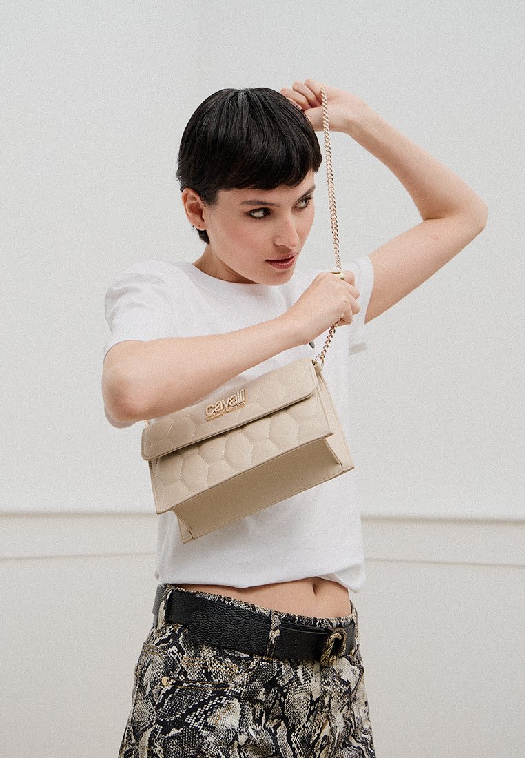 Bolso acolchado beige con un patrón hexagonal, herrajes dorados y una correa de cadena, sostenido por una persona con una camisa blanca y pantalones estampados.