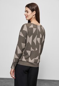 Pull à manches longues en vert olive avec des motifs géométriques beiges. Tissu doux avec une coupe décontractée et un large col, comportant un détail de couture au dos.