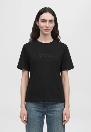 Modèle portant un T-shirt BOSS noir à manches courtes avec un logo discret et un jean bleu, sur un fond uni.