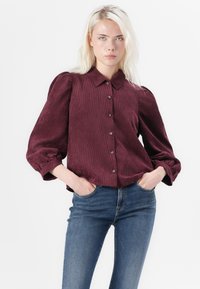Burgundi värvi korduroy särk, millel on krae, nööbistatud ees ja puhvärv. Kandmine siniste teksadega, mis näitavad lõdva istuvuse ja tekstuurset materjali.