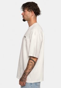 Tricou oversized din bumbac de culoare crem, cu mâneci scurte, având un mic logo imprimat pe piept. Tatuaje vizibile pe braț.