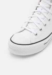 Zapatilla de lona blanca con punta redonda, suela de goma negra y agarre texturizado. Presenta ojales de metal plateado y cordones blancos.
