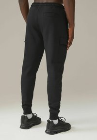 Pantalones deportivos negros con un ajuste ajustado, cinturilla elástica, dos bolsillos laterales tipo cargo y puños de canalé, combinados con zapatillas deportivas negras y calcetines blancos.