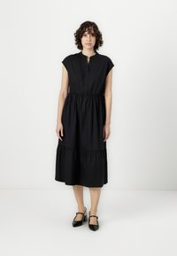 Black cap-sleeve dress with a stand collar, gathered waist, and tiered skirt. Smooth texture with a flowing design. Worn with black heels.