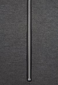 Grå tyg jacka med dragkedja framtill, med en central silverfärgad dragkedja, mjuk textur och kontrasterande svart kant längs dragkedjan.