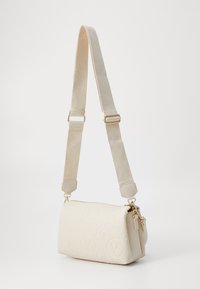 Beige crossbody bag features embossed logo pattern, gold hardware accents, smooth texture, and an adjustable strap. Zipper closure at the top.