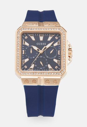 Montre-bracelet carrée avec un boîtier en or rose, un cadran bleu et un bracelet en silicone bleu. Présente des accents en cristal et trois sous-cadrans pour la mesure du temps.