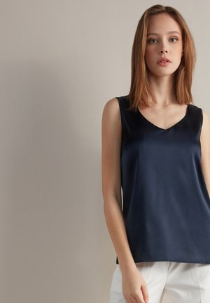 Haut sans manches en satin bleu marine avec un décolleté en V ; texture lisse ; coupe décontractée ; associé à un pantalon de couleur claire.