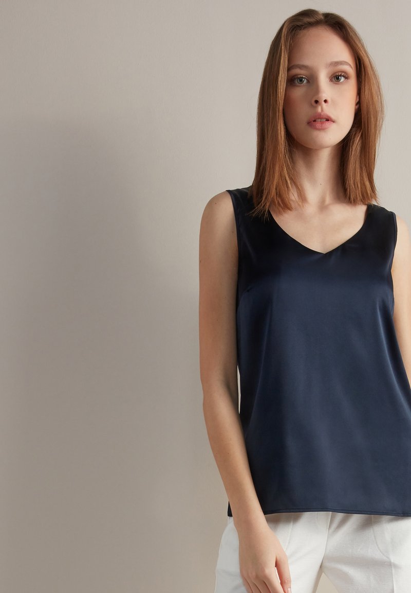 Granatowa satynowa bluzka bez rękawów z dekoltem w kształcie litery V; gładka faktura; luźny krój; zestawiona z jasnymi spodniami.