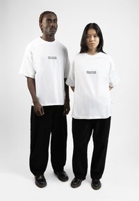 Dos individuos están uno al lado del otro, vistiendo camisetas blancas de gran tamaño con un pequeño gráfico en el centro, combinadas con pantalones de pana negros.