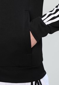 Černá mikina s vysokým límcem, zdobená bílými pruhy na rukávech a boční kapsou. Měkká, texturovaná tkanina.