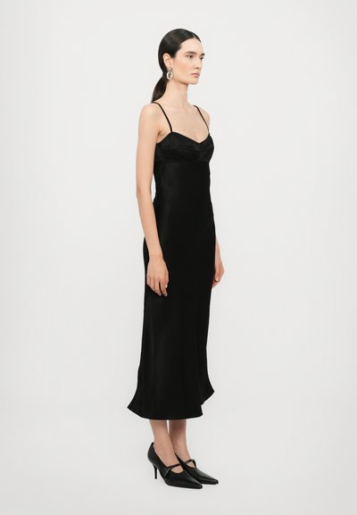 Vestido de satén negro sin mangas, con un corpiño ajustado y un dobladillo acampanado. Combinado con tacones negros de punta para un look estilizado.