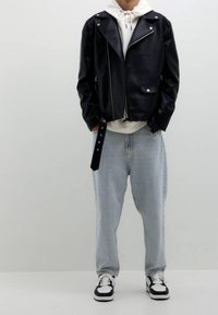 Chaqueta de cuero negra estilo motorista con herrajes plateados, cierre de cremallera y bolsillos laterales sobre una sudadera con capucha blanca, combinada con vaqueros anchos azul claro y zapatillas.