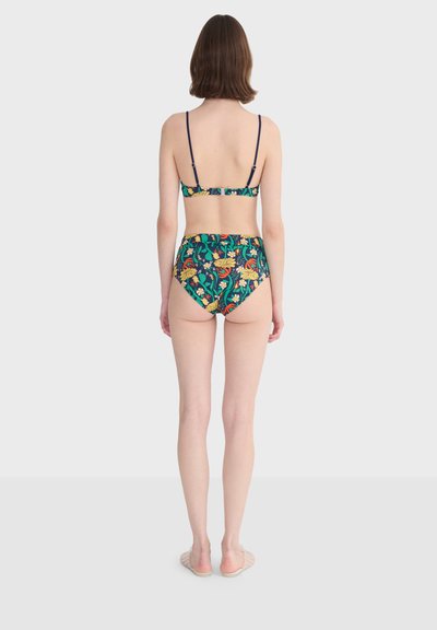 Femme debout de dos, portant un bikini taille haute à motif floral foncé et des sandales plates sur un fond clair uni.