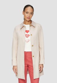 Lichtbeige, lange mouwen jas met een kraag en gouden knopen, gedragen over een wit shirt met rode hartafdrukken en gecombineerd met koraalkleurige broek.