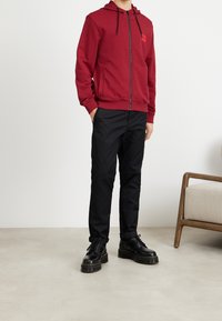 Červená mikina na zip s logem na přední straně, doplněná černými slim-fit kalhotami a černými šněrovacími botami s tlustou podrážkou.