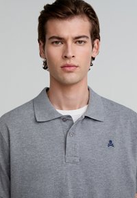 Grijze polo met textuurstof, voorzien van een kraag, twee knopen en een klein blauw doodskoplogo op de linkerborst.