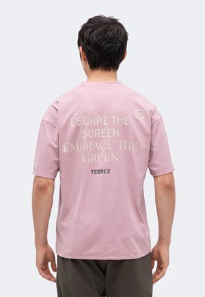 Mand med kort mørkt hår iført en pink t-shirt med teksten "FLUGT FRA SKÆRMEN, OMARM GRØNT TERREX" på ryggen.