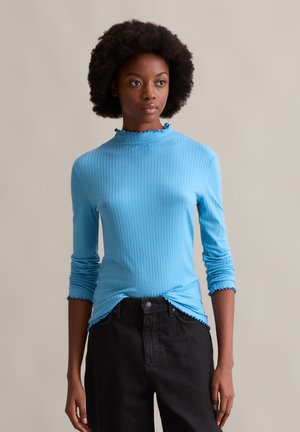 Light blue ribbed long-sleeve top with a high neckline and ruffled trim, paired with black trousers. Smooth texture, fitted design.