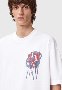 Hvit bomulls t-skjorte med et dristig grafisk design i rødt og blått, med dryppende tekst innenfor et sirkulært mønster på fronten.