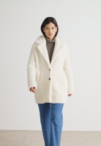 White faux fur coat with a collar, two front pockets, and button closure. Paired with light blue straight-leg jeans.