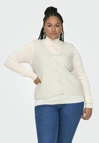 Elfenbensfärgad stickad väst med texturerad tyg och fem knappar, lager på lager över en krämfärgad höghalser. Bärs med blå jeans och har en avslappnad passform.