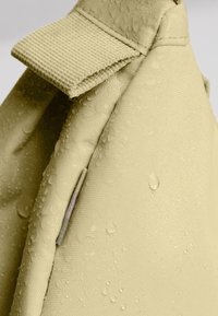 Tejido impermeable en amarillo-verde con superficie texturizada. Presenta una pestaña y gotas de agua visibles, destacando su calidad resistente al agua.