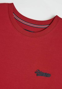 Czerwona bawełniana koszulka z okrągłym dekoltem. Posiada czarne haftowane logo oraz paskowany wewnętrzny kołnierzyk w kolorach czerwonym, białym i niebieskim. Gładka tekstura.