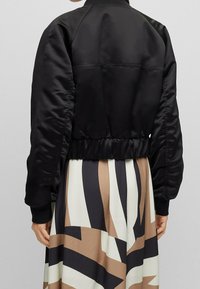 Svart satin bomberjacka med en kortad design, elastisk midja och ribbade ärmslut. Slät textur med en minimalistisk stil.