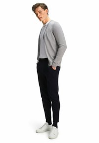 Cardigan grigio chiaro con chiusura a bottoni, indossato sopra una camicia bianca, abbinato a pantaloni scuri e sneakers bianche, creando un look a strati.