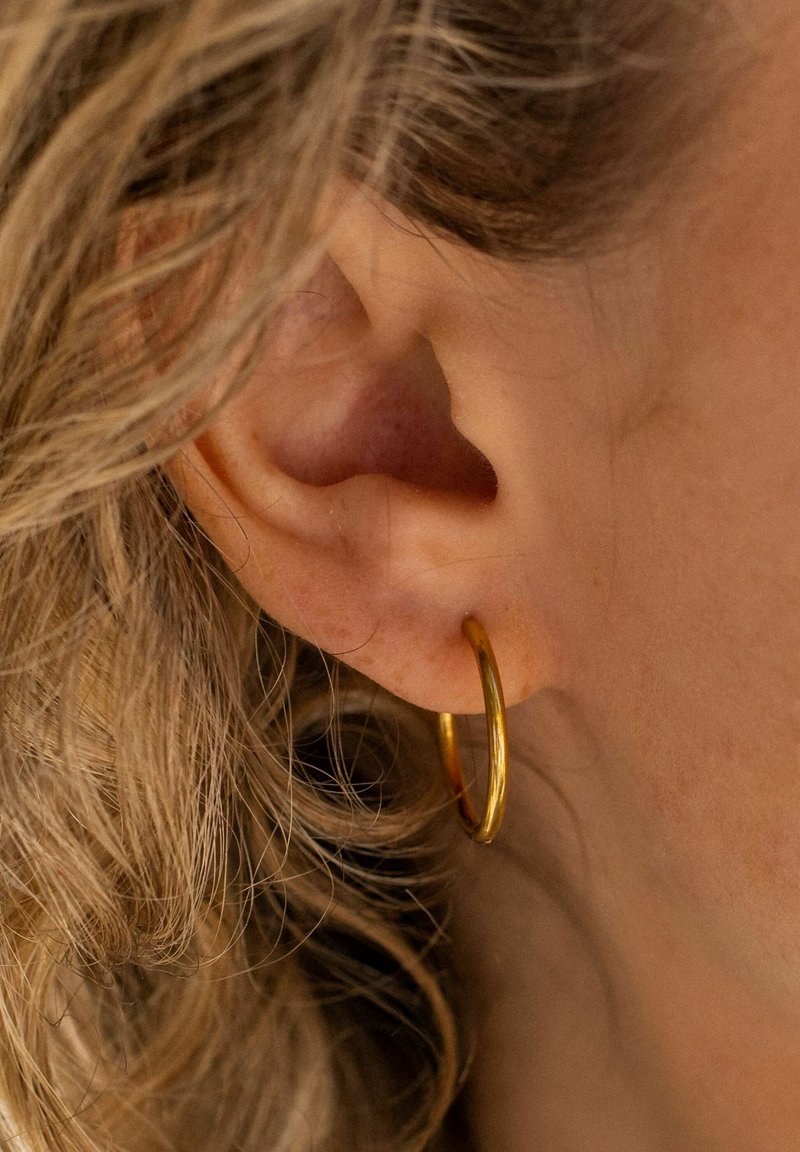 Goldfarbene Metall-Hoop-Ohrringe, mittelgroß, am Ohrläppchen getragen, mit einer glatten, glänzenden Oberfläche und einem dünnen, kreisförmigen Design. Lockiges Haar im Hintergrund.