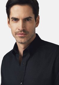 Czarna koszula z guzikami, z wyraźnym kołnierzem, teksturowanym materiałem i małą kieszonką na lewej piersi z subtelnym detalem logo.