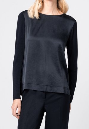 Vrouw die een zwarte longsleeve draagt met een glad satijnen voorpaneel en bijpassende zwarte broek tegen een effen achtergrond.