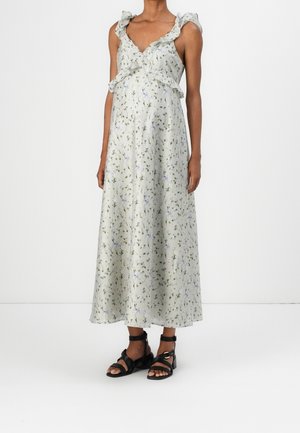 Kobieta w jasnej, kwiecistej, bezrękawnikowej sukience maxi z falbanami na ramiączkach i czarnych sandałach na pasku przy kostce, stojąca na jednolitym tle.