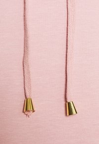 Sudadera de color rosa claro con cordones trenzados que presentan terminales de metal dorado, sobre un fondo de tela suave.