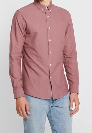Chemise boutonnée rose clair avec un col, boutons blancs et un petit logo brodé. Le matériau semble être du coton, de conception ajustée, à manches longues.