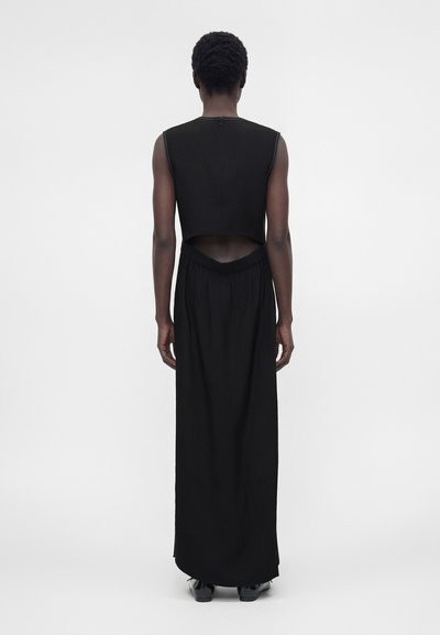 Άτομο με κοντά σγουρά μαλλιά στέκεται πλάτη, φορώντας αμάνικη μαύρη μπλούζα με άνοιγμα στην πλάτη και μακριά μαύρη φούστα με μαύρα παπούτσια.