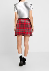 Roter karierten Rock mit Falten, kombiniert mit einem weißen gestreiften Kurzarmhemd. Schwarze Knöchelstiefel mit dicken Sohlen werden getragen.