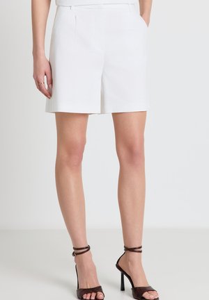 Donna con pantaloncini bianchi su misura con tasche laterali e sandali marroni con tacco alto e cinturino alla caviglia, in piedi davanti a uno sfondo semplice.